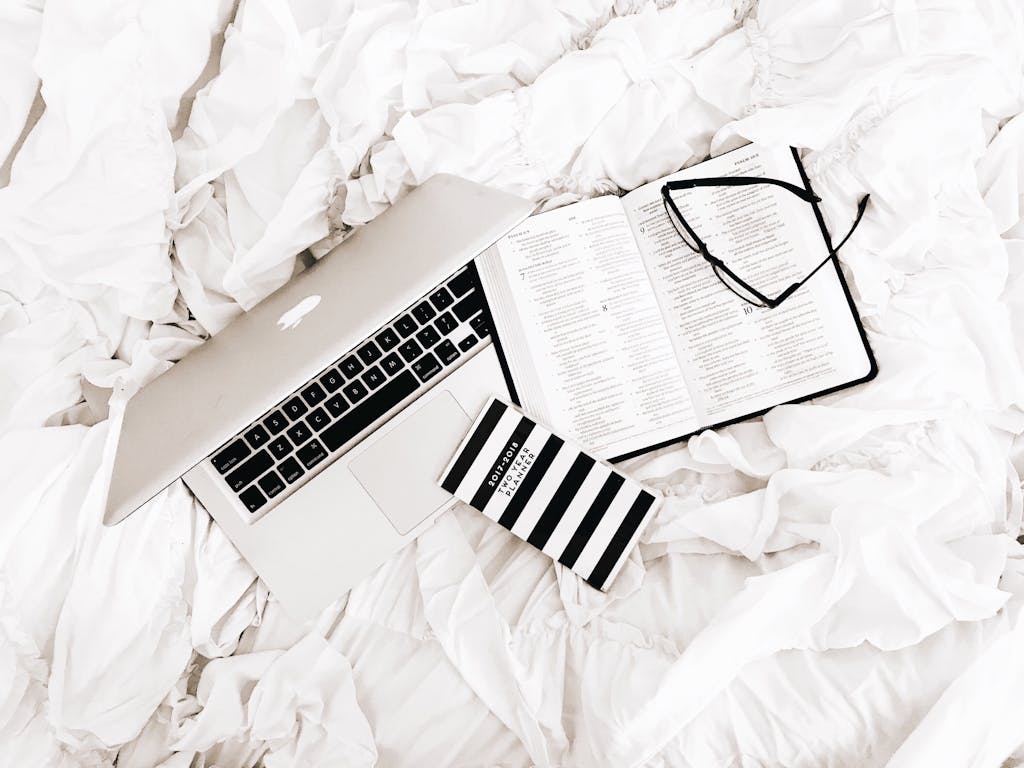 A flat lay of a laptop, open book, and reading glasses on a white bed. Perfect for productivity and relaxation themes.